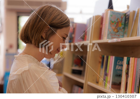 Woman browsing children's books in a cozy library while holding a baby in a quiet afternoon setting 130410307