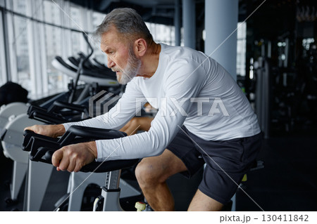 Elderly man riding an exercise bike in a bustling gym 130411842
