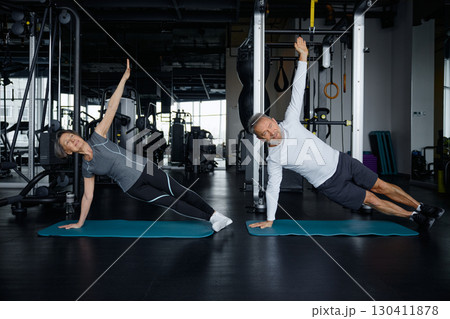 Mature man and a woman are performing side plank exercises at a gym facility Mature man and a woman are performing side plank exercises at a gym facility 130411878