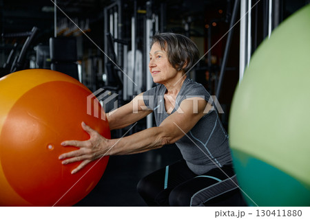 An older woman is squatting with an orange ball in a gym An older woman is squatting with an orange ball in a gym 130411880