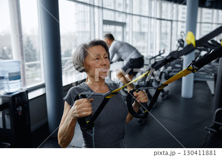 Mature woman is exercising on a suspension trainer in a gym 130411881