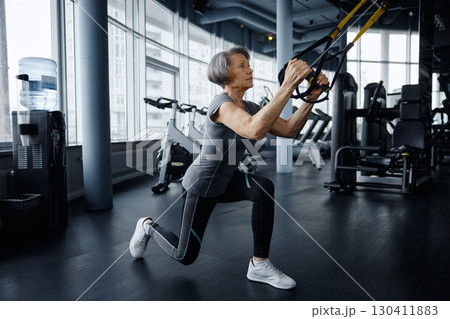 An elderly woman is exercising on a suspension trainer in a gym 130411883