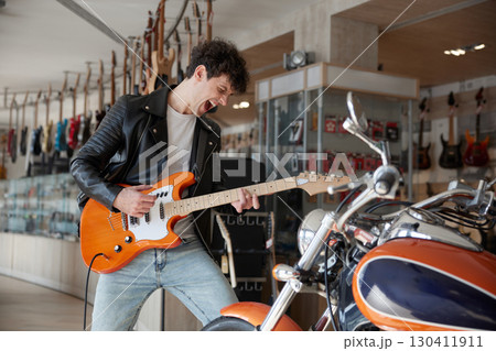 Expressive man in a leather jacket is playing guitar in a store 130411911