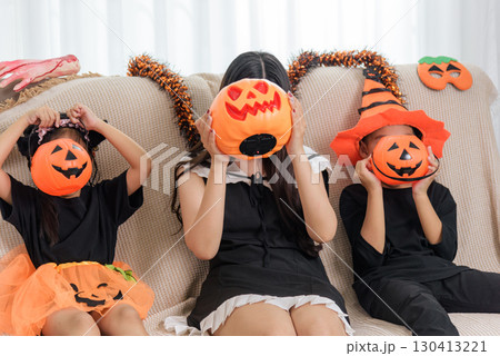 Three people celebrate the Halloween holiday season on a sofa, holding bright plastic pumpkin containers over their faces as part of festive dress up. 130413221