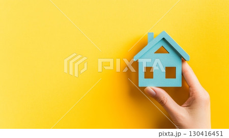 Hand holds a small blue house against a vibrant yellow backdrop, representing real estate investment, homeownership, and the mortgage process 130416451