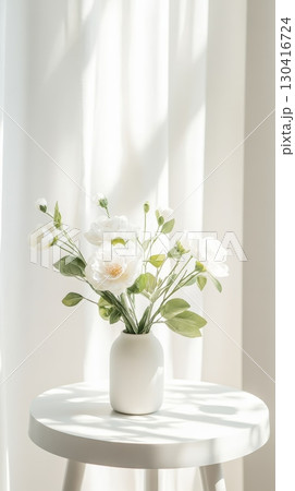 Sunlight streams through sheer white curtains illuminating a white vase filled with white flowers sitting on a small, round, white table, creating a serene and minimalist scene 130416724