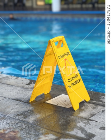 Yellow caution sign on wet poolside surface near swimming pool, warning of cleaning in progress and slippery floor hazard for safety awareness. 130417971