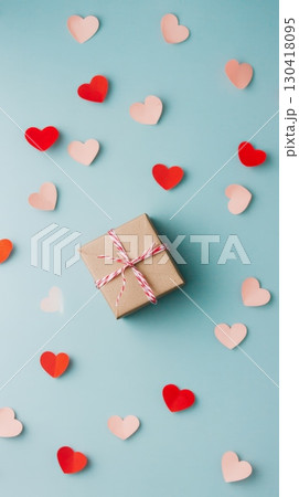 Small wrapped gift box adorned with red and white ribbon, surrounded by red and pink paper hearts, creating a romantic vibe on a pastel blue backdrop 130418095