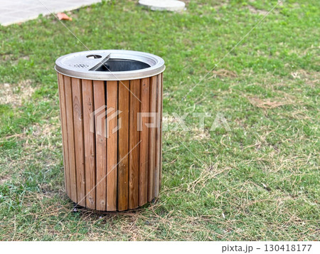 Wooden trash bin with metal lid stands on grass in public park, copy space. Ecology, sustainability, and waste management reflecting cleanliness, urban service and environmental care outdoors. Wooden trash bin with metal lid stands on grass in public park, copy space. Ecology, sustainability, and waste management reflecting cleanliness, urban service and environmental care outdoors. 130418177