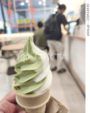 a soft serve ice cream cone with green tea and vanilla swirl, held indoors at a modern cafe with blurred background. a soft serve ice cream cone with green tea and vanilla swirl, held indoors at a modern cafe with blurred background. 130418430