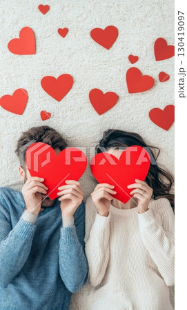 Young couple lying on carpet hiding their faces with red paper hearts surrounded by other small hearts celebrating valentine's day, love, and spring Young couple lying on carpet hiding their faces with red paper hearts surrounded by other small hearts celebrating valentine's day, love, and spring 130419097