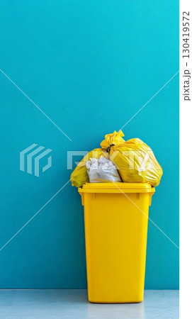 Overflowing yellow trash can with garbage bags against a vibrant teal wall, highlighting the importance of proper waste disposal and recycling practices for environmental sustainability 130419572