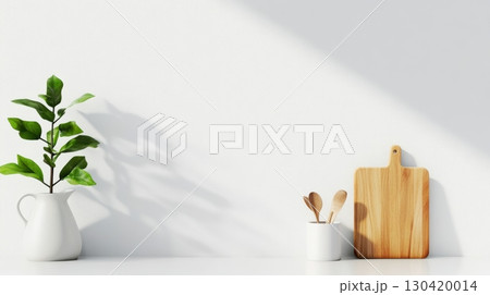 Minimalist kitchen counter featuring a plant in a white vase, a cutting board, and wooden spoons in a white container, bathed in sunlight casting shadows on a white wall 130420014