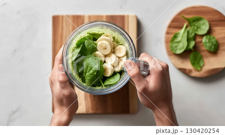 Chef is adding banana slices to a blender glass already containing spinach and a green smoothie base, getting ready to create a healthy and refreshing beverage 130420254