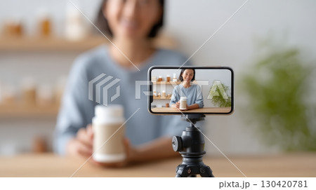 Asian female pharmacist is recording a video blog about vitamins and supplements, holding a white plastic bottle with blank label while sitting at desk in front of smartphone fixed on tripod Asian female pharmacist is recording a video blog about vitamins and supplements, holding a white plastic bottle with blank label while sitting at desk in front of smartphone fixed on tripod 130420781
