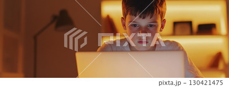 Focused boy studying at night in his bedroom, illuminated by the soft glow of the laptop screen, diligently completing homework and enhancing his learning experience 130421475