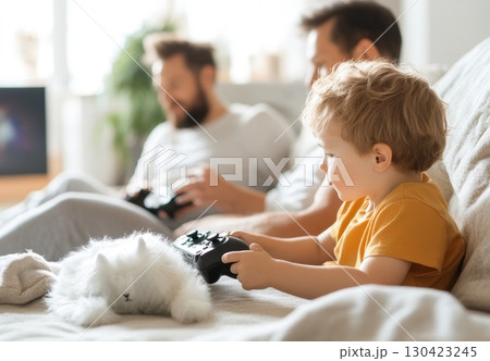 A toddler joyfully plays a video game while his fathers relax nearby, enjoying a cozy family moment at home A toddler joyfully plays a video game while his fathers relax nearby, enjoying a cozy family moment at home 130423245