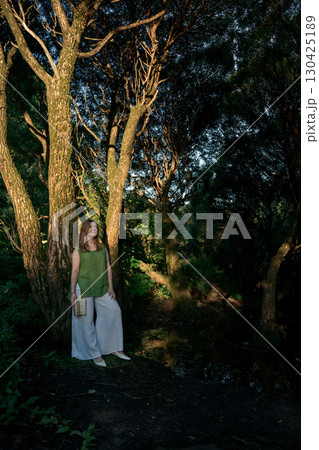 Woman in a green top and white trousers stands with a book in hand, leaning against a tree in Izmailovsky Park, Moscow 130425189