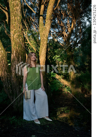 Woman in a green top and white trousers stands with a book in hand, leaning against a tree in Izmailovsky Park, Moscow 130425226