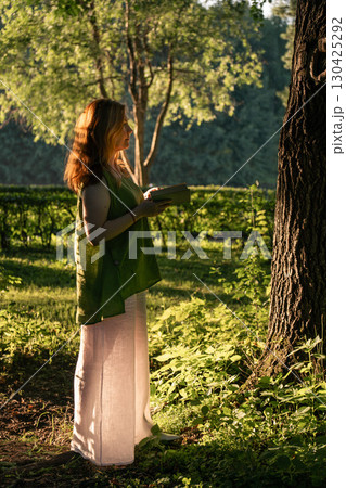 Woman in a green linen top and white trousers stands by a tree with a book in her hands, bathed in golden evening light in Izmailovsky Park, Moscow 130425292