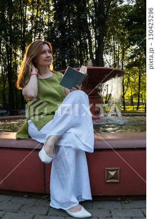 A young woman sits by a fountain in a park, holding an open book and gazing thoughtfully into the distance 130425296