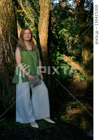 Woman in a green linen top and white trousers leans against a tree while holding an open book in Izmailovsky Park, Moscow Woman in a green linen top and white trousers leans against a tree while holding an open book in Izmailovsky Park, Moscow 130425308