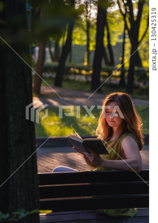 A woman sits on a park bench at sunset, reading an old book while warm golden light highlights her hair and face A woman sits on a park bench at sunset, reading an old book while warm golden light highlights her hair and face 130425319