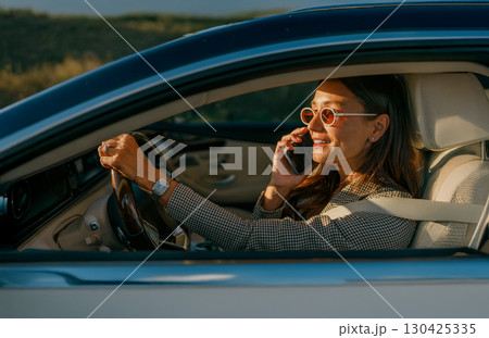 A Stylish Woman is Driving a Car while Engaged in a Phone Call during a Beautiful Day 130425335