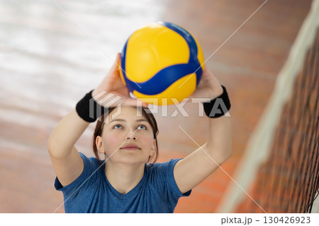 Young girl setting the ball over the net before hitting it. Pupil of high school practicing volleyball skills, PE lesson at school in the gym Young girl setting the ball over the net before hitting it. Pupil of high school practicing volleyball skills, PE lesson at school in the gym 130426923