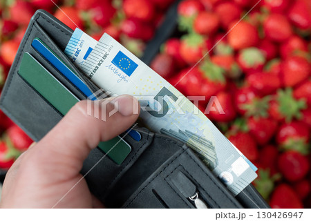 Wallet with euro money and credit cards in hand of a man, fresh strawberries on the background, close up. Concept of organic farming, earning money in the countryside, seasonal purchase Wallet with euro money and credit cards in hand of a man, fresh strawberries on the background, close up. Concept of organic farming, earning money in the countryside, seasonal purchase 130426947