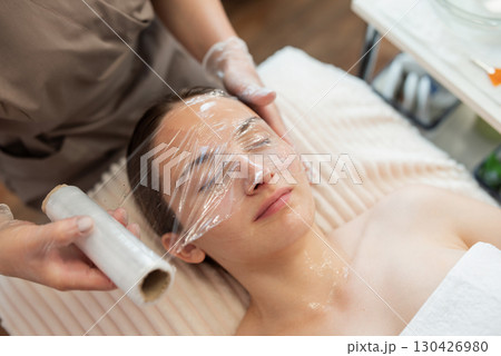 Covering face of a client in plastic material after applying gel for for cold steaming of the skin before cleaning. Beauty cosmetology clinic, cleaning skin and pores on face Covering face of a client in plastic material after applying gel for for cold steaming of the skin before cleaning. Beauty cosmetology clinic, cleaning skin and pores on face 130426980