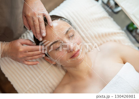Face of a client of cosmetology clinic covered in plastic material after applying gel for pores to open and better ultrasound or mechanical cleaning. Cleaning of the face of a client in beauty salon 130426981