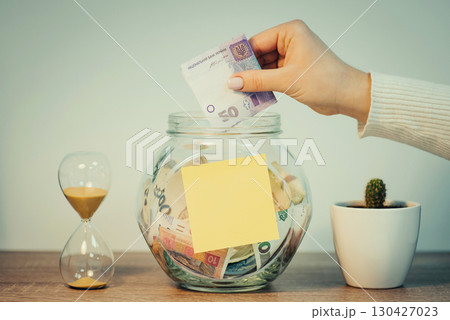 Ukrainian person saving up money for a big purchase. Girl puts 50 hryvnias banknote to the glass jar with blank paper for inscription, toned photo Ukrainian person saving up money for a big purchase. Girl puts 50 hryvnias banknote to the glass jar with blank paper for inscription, toned photo 130427023