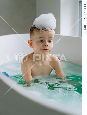 Sunlight pours through the floor-to-ceiling windows as the boy relaxes in a pristine white bathtub, the warm glow reflecting off the glossy tiles of the spacious bathroom. High quality photo Sunlight pours through the floor-to-ceiling windows as the boy relaxes in a pristine white bathtub, the warm glow reflecting off the glossy tiles of the spacious bathroom. High quality photo 130427544