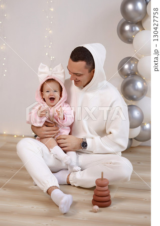 Father and baby in hoodies sitting on wooden floor at birthday party with balloons and white wall Father and baby in hoodies sitting on wooden floor at birthday party with balloons and white wall 130427758