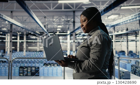 IT expert monitors server farm activity using laptop while walking in facility. African american woman in data center reviews rigs tests results to ensure systems are working correctly, camera A 130428690
