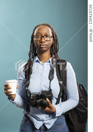Female photographer standing with camera and disposable cup in her hands. African American female model poses confidently, wearing blue shirt, with DSLR around neck and holding coffee. 130428726