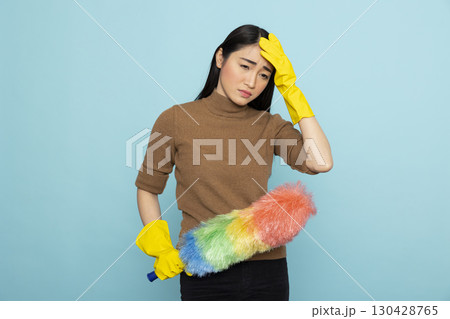 Portrait of overwhelmed cleaning service staff holding feather duster, experiencing job fatigue. Asian housemaid in yellow rubber gloves expressing exhaustion, standing isolated over blue background. Portrait of overwhelmed cleaning service staff holding feather duster, experiencing job fatigue. Asian housemaid in yellow rubber gloves expressing exhaustion, standing isolated over blue background. 130428765