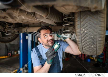 Professional mechanic inspecting car's undercarriage with flashlight in a repair shop 130428788