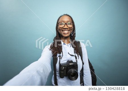 Pov of african american woman holding camera to record herself, grinning as she creates content in studio with dslr around her neck. Black female photographer smiling and taking a self portrait. Pov of african american woman holding camera to record herself, grinning as she creates content in studio with dslr around her neck. Black female photographer smiling and taking a self portrait. 130428818