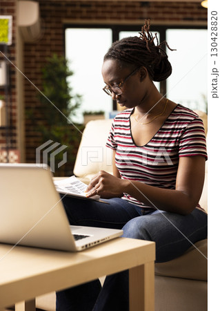 African american woman working from home, sitting on couch with clipboard in hand, reading through paperwork. Female freelancer with glasses flipping printed documents, planning and organizing tasks. 130428820