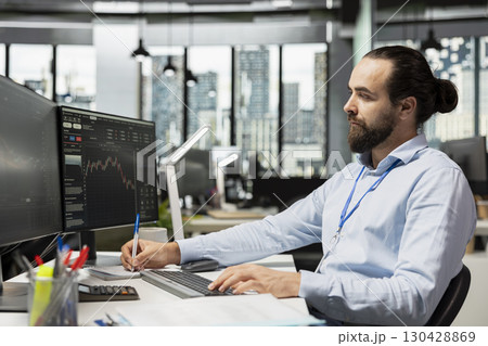 Risk analyst in hedge fund office taking notes while analyzing candlestick charts and price graphs on screen. Brokerage company worker writing analysis of market trends and trading strategies Risk analyst in hedge fund office taking notes while analyzing candlestick charts and price graphs on screen. Brokerage company worker writing analysis of market trends and trading strategies 130428869