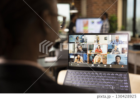 Rearview closeup of black businesswoman attending videoconference call with multiethnic colleagues working remotely. The focus is selectively on laptop screen displaying an ongoing virtual meeting. Rearview closeup of black businesswoman attending videoconference call with multiethnic colleagues working remotely. The focus is selectively on laptop screen displaying an ongoing virtual meeting. 130428925