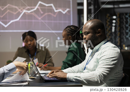 Diverse employees examining annual data reporting in a firm meeting at night, looking into market trends on analytics to facilitate global business development. Work team planning. 130428926