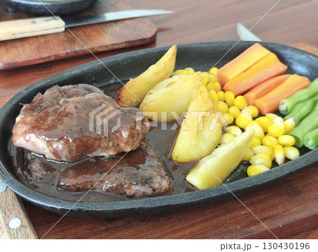 Sizzling hot plate of steak with rich brown sauce, served with potato wedges, corn, carrots, and green beans on a cast iron skillet. 130430196