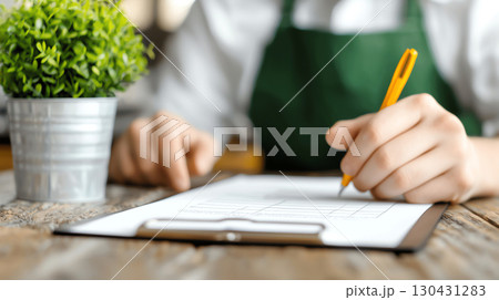 Closeup barista coffee shop manager owner in green apron writes prices taxes record calculation profit and loss on clipboard at wooden table with potted plant nearby, focusing on details 130431283