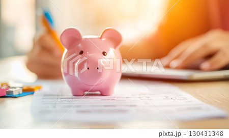 pink piggy bank sits on table with documents, as person reviews financial information in background, highlighting concept of saving and budgeting 130431348