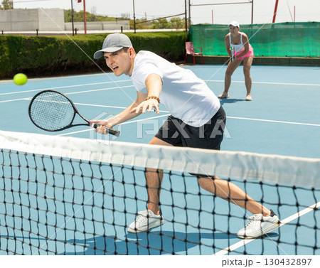 Portrait of young guy playing tennis on open court in summer, swinging racket to return ball over net. Sportsman ready to hit volley 130432897