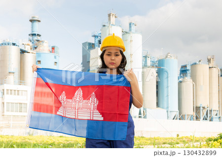 Sad young woman in helmet holding flag of Cambodia against background of factory 130432899