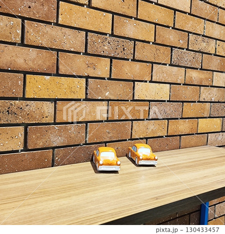 Two small, orange toy cars rest on a light wooden surface near the brick wall  130433457